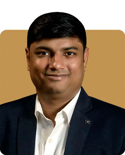 A man smiling, wearing a dark blazer over a white shirt, against a warm-colored background.