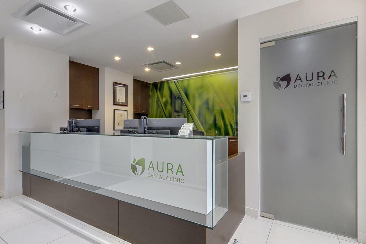 A modern dental clinic reception area with a glass counter displaying the name "Aura Dental Clinic," featuring two computer stations and a green-themed backdrop.