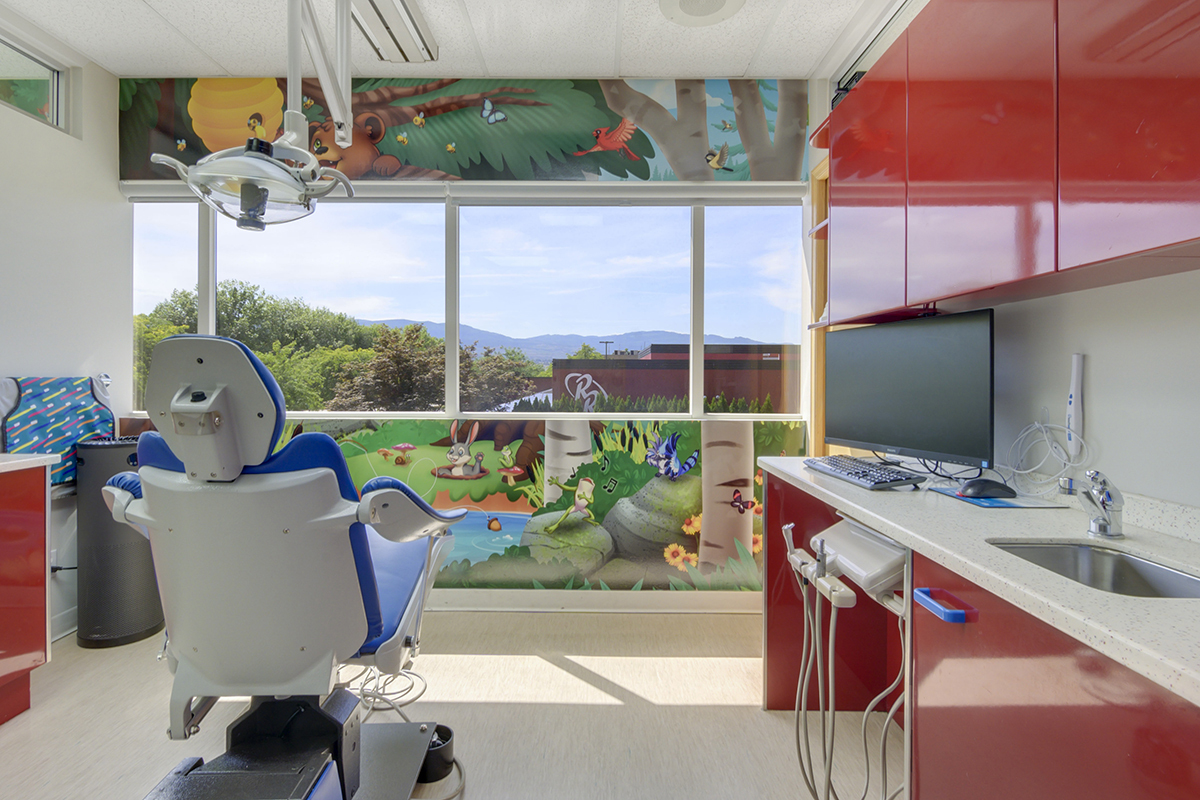 A colorful pediatric dental office featuring a treatment chair, bright red cabinetry, and a mural with cartoon animals and nature.