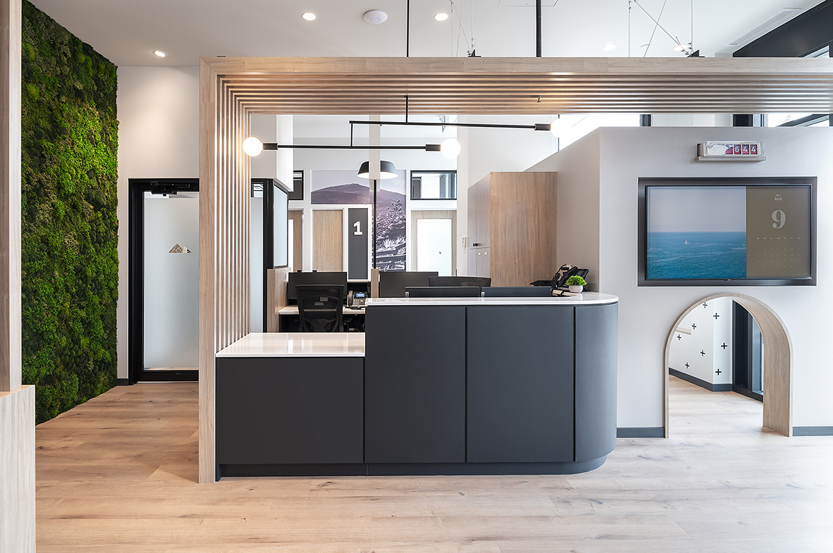 A modern office interior featuring a reception area with a curved black desk, a moss wall, wooden accents, and open workstations.