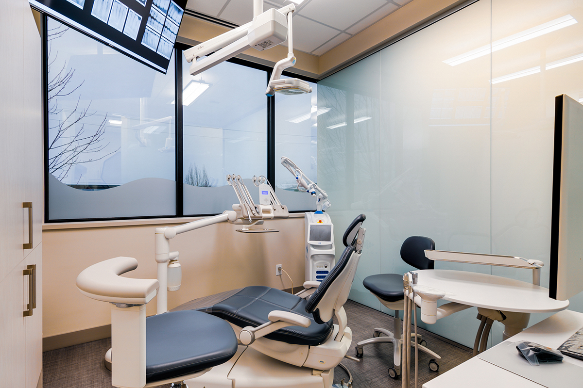 A modern dental office with a dental chair, equipment, a monitor displaying x-rays, and large windows.
