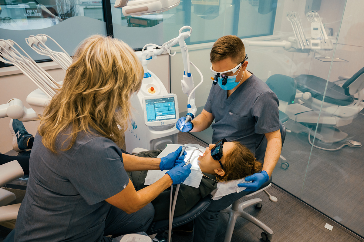A dental procedure is taking place, with a patient reclining in a dental chair while two dental professionals work on her teeth.