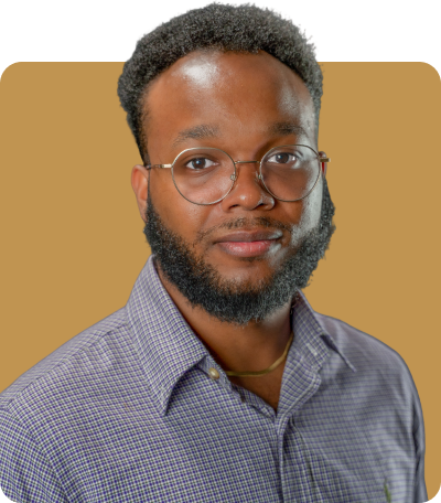 Head-and-shoulders portrait of a young man with a short beard and curly hair, wearing round glasses and a checkered button-down shirt, posed against a neutral light brown background
