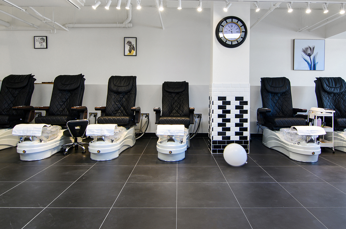 A modern nail salon featuring black leather pedicure chairs, pedicure basins, a wall clock, and decorative framed artwork.