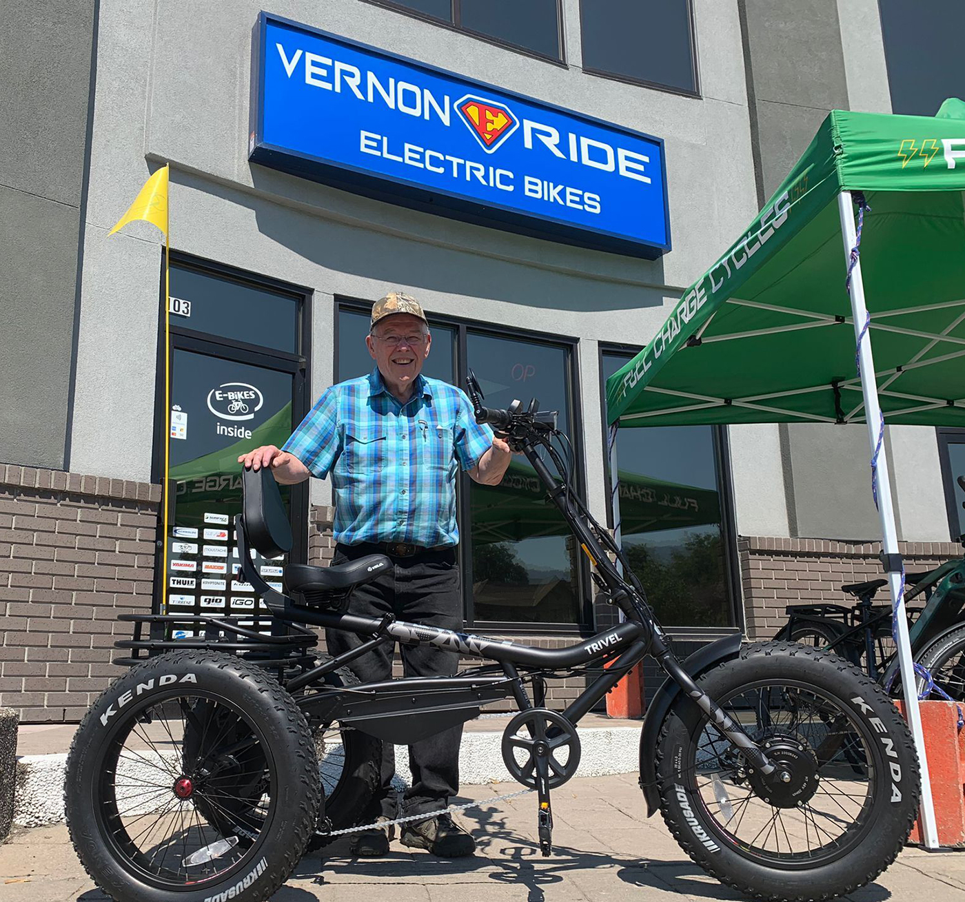 An older man in a plaid shirt stands next to an electric bike outside a store named "Vernon Ride Electric Bikes." The store has a green tent and signage promoting electric bikes.