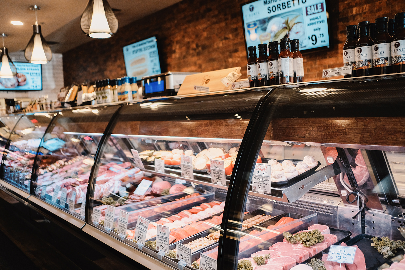 A glass display case filled with various meats, including fish and cuts of beef, pork, and seafood, with signage indicating prices and specials. Above, bottles of sauce and menus are visible.