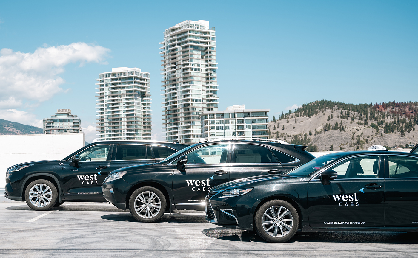 Three black taxi vehicles with "west CABS" branding parked in a row, with modern buildings and mountains in the background.