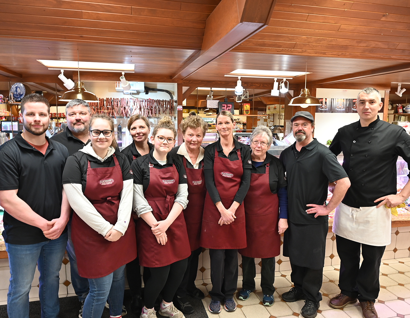 A group of ten people standing together in a store, all wearing black shirts and maroon aprons, with a meat display in the background.