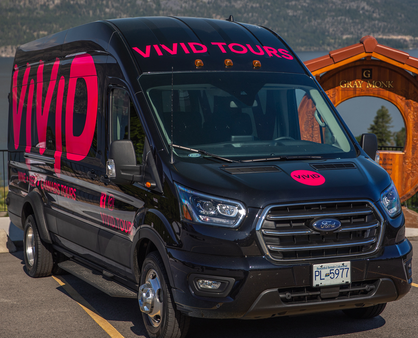 A black van with "VIVID TOURS" printed in large pink letters, parked near a wooden sign that reads "GRAY MONK."