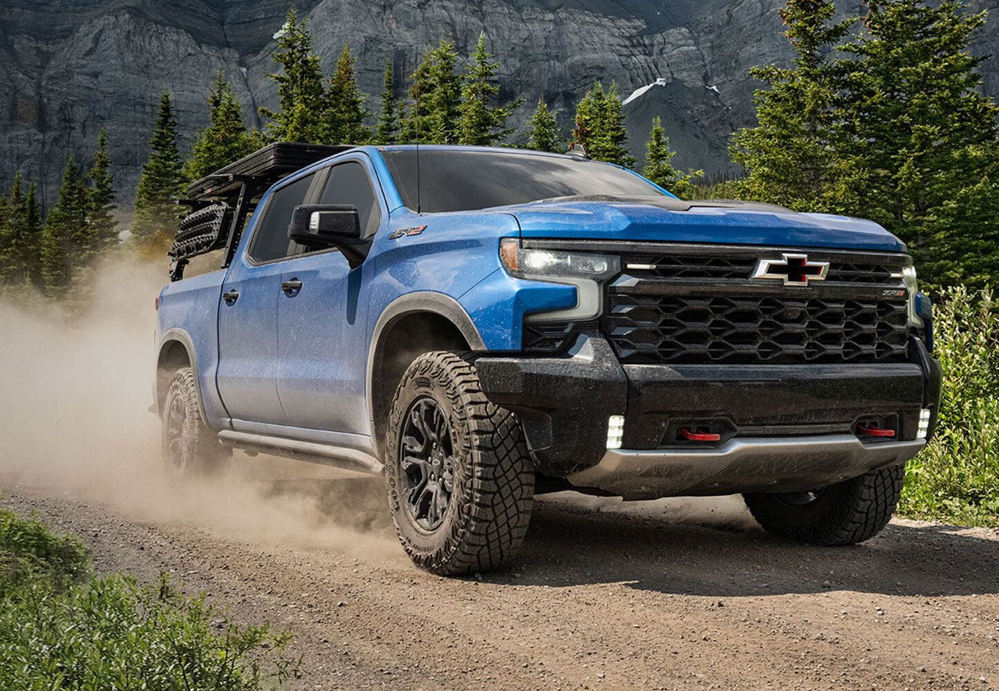 A blue Chevrolet pickup truck driving on a dirt road surrounded by trees and mountains. Dust is being kicked up behind the vehicle.