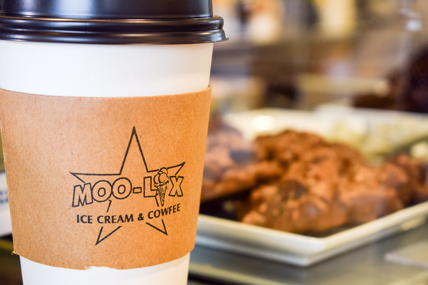A coffee cup with a paper sleeve featuring the logo "MOO-LIX ICE CREAM & COWFEE," with cookies blurred in the background.