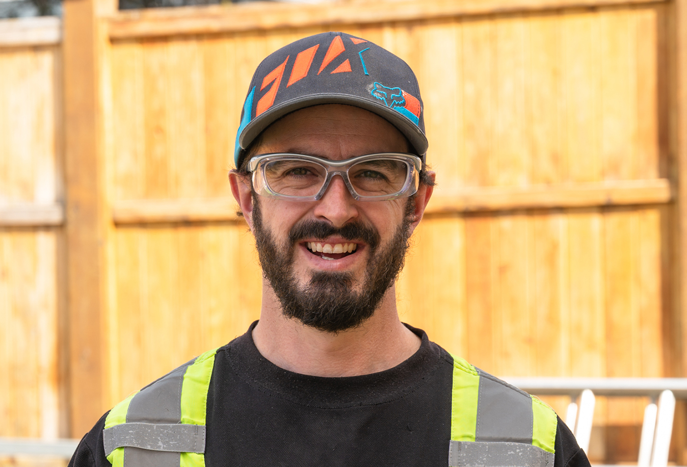 A man wearing safety glasses and a cap, smiling, in front of a wooden fence. He has a beard and is in a work shirt with reflective stripes.