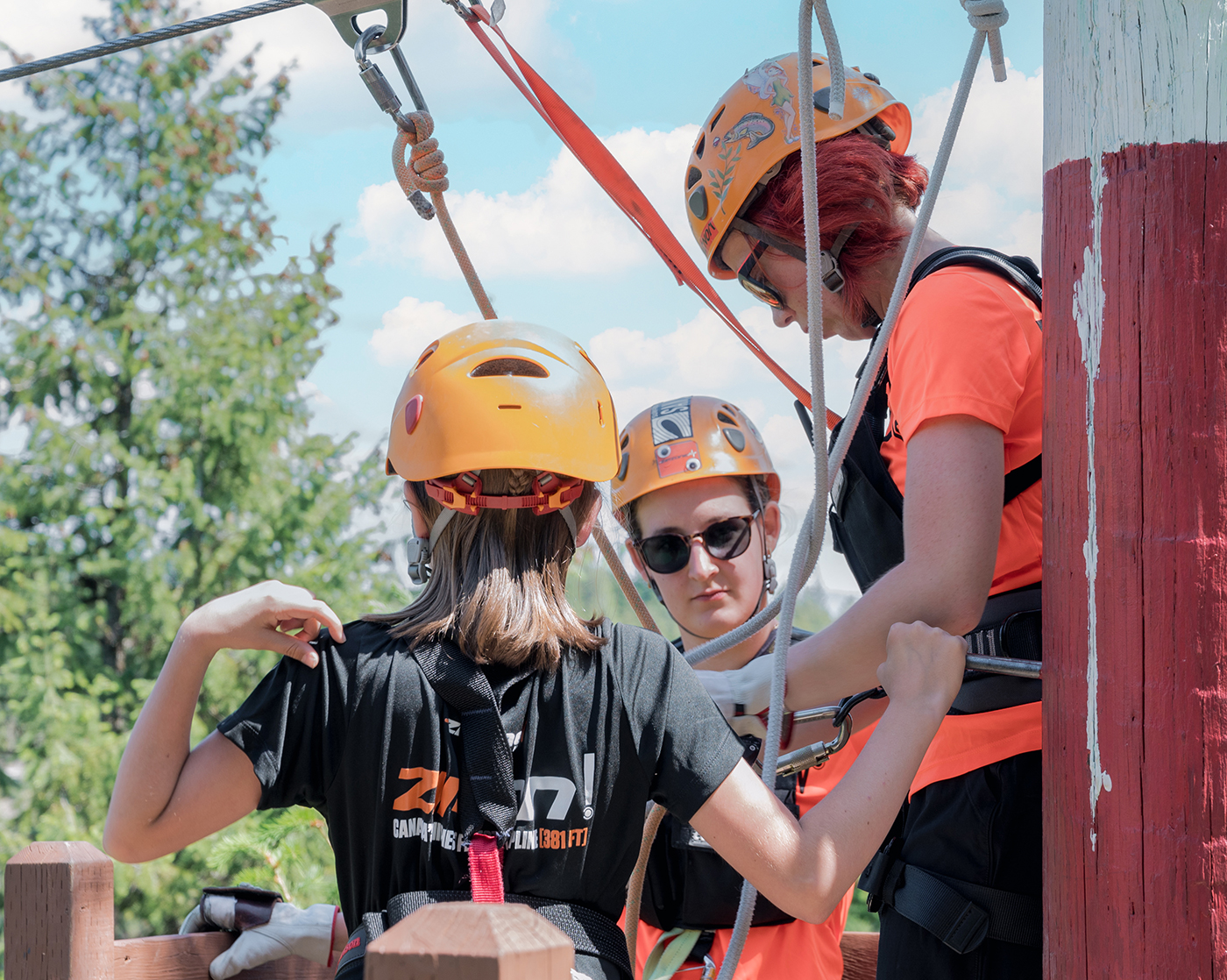 Three individuals wearing helmets and safety gear are preparing for an outdoor activity, possibly zip-lining.