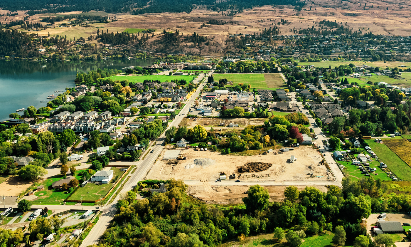 Aerial view of a residential area near a lake, featuring houses, roads, and some construction sites amidst greenery and hills.