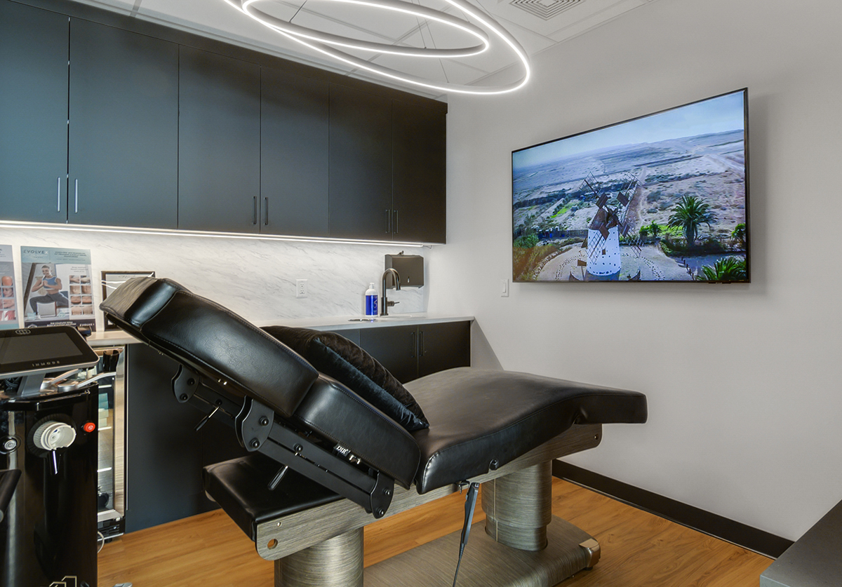 A modern treatment room featuring a black reclining chair, a large wall-mounted TV displaying a scenic landscape, and dark cabinetry.