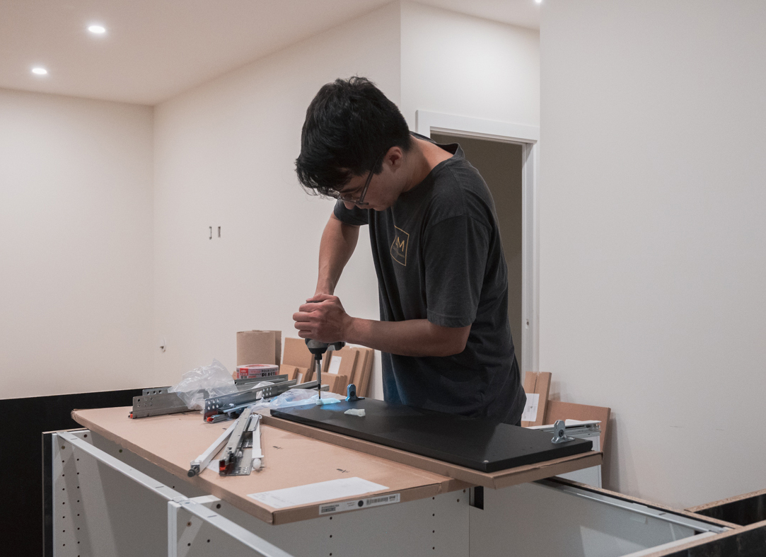 A person is assembling furniture using a screwdriver on a tabletop, surrounded by tools and materials in a room.