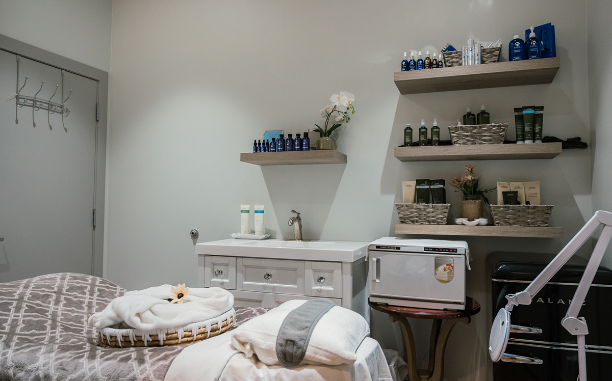 A soothing spa room featuring a massage table with towels, shelves displaying skincare products, a small refrigerator, and a treatment cart.