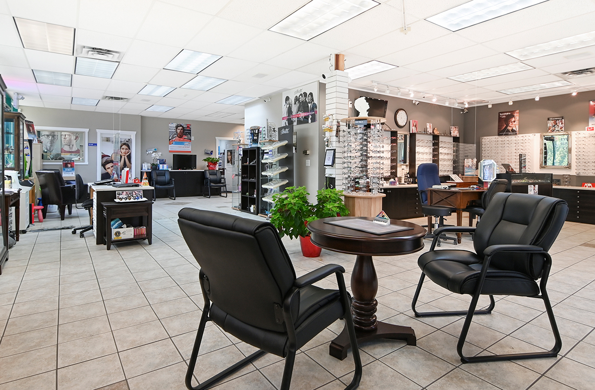 A spacious eyewear store featuring display shelves of glasses, a reception area with a desk, black chairs, and a variety of promotional materials on the walls.