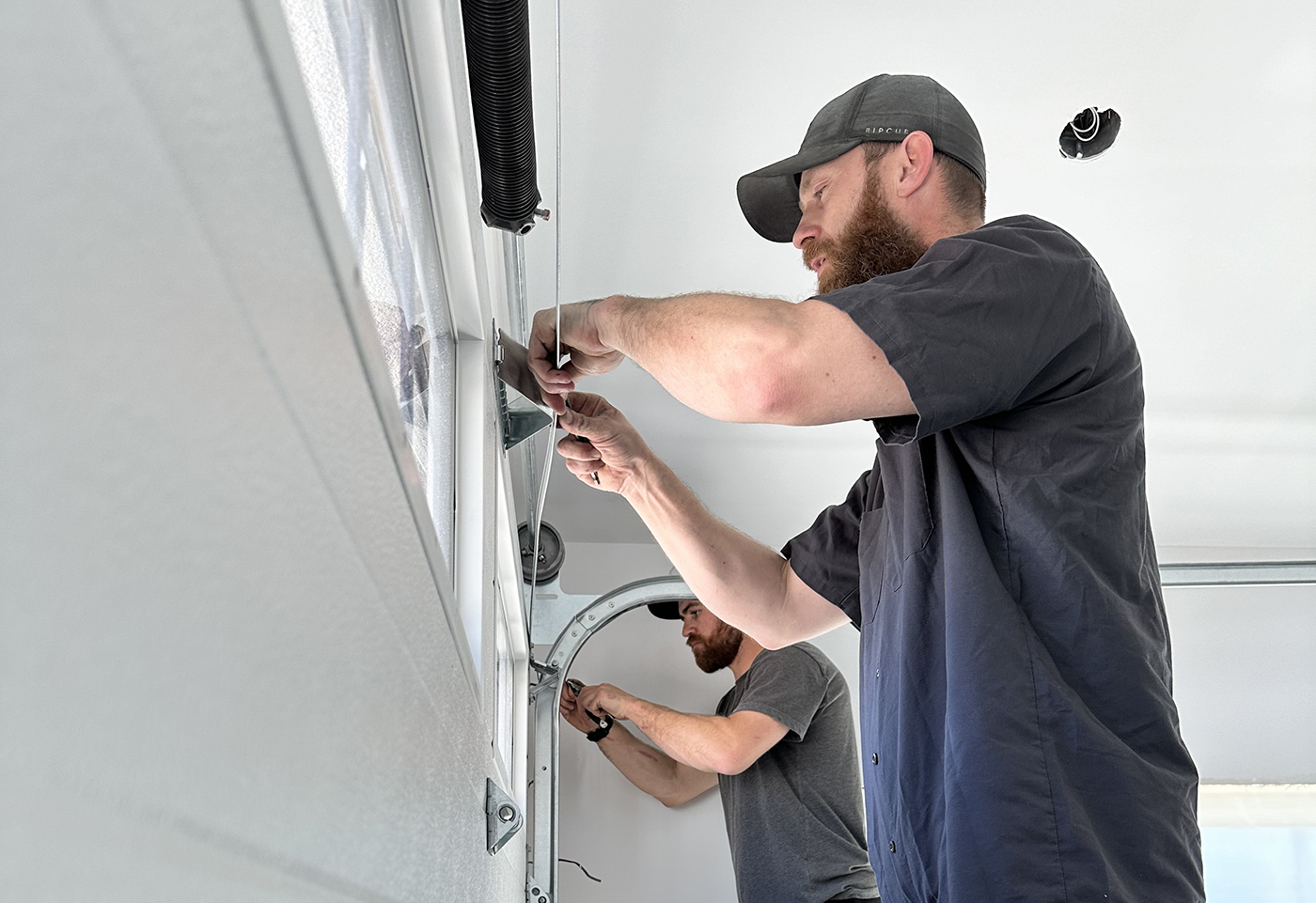 Two men are working on installing or repairing equipment in a garage, focusing on a mechanism mounted on the wall.