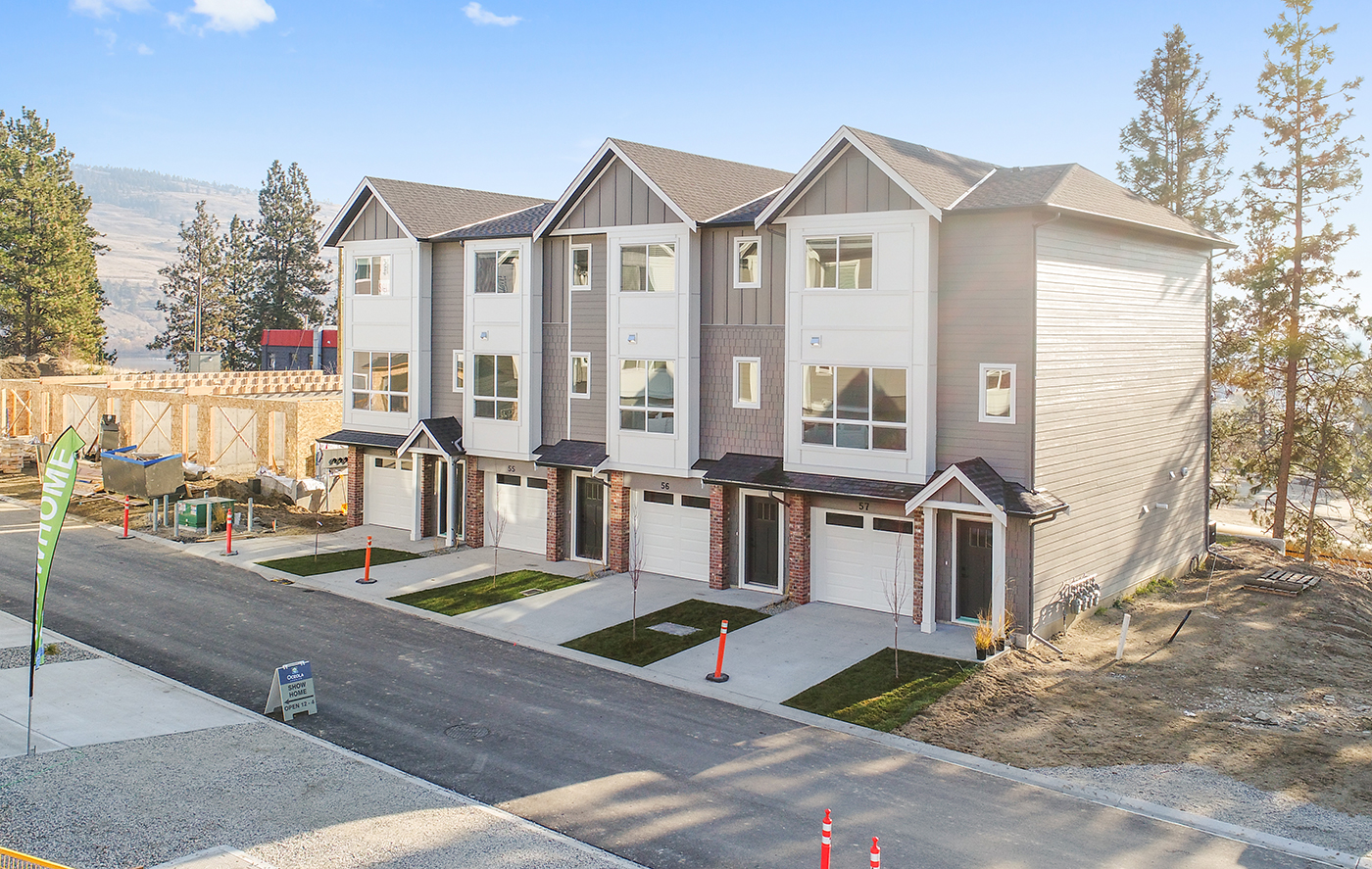A row of modern townhouses with gray and white exteriors, featuring small front yards and driveways. A construction site is visible in the background, along with traffic cones and a 'Show Home' sign.