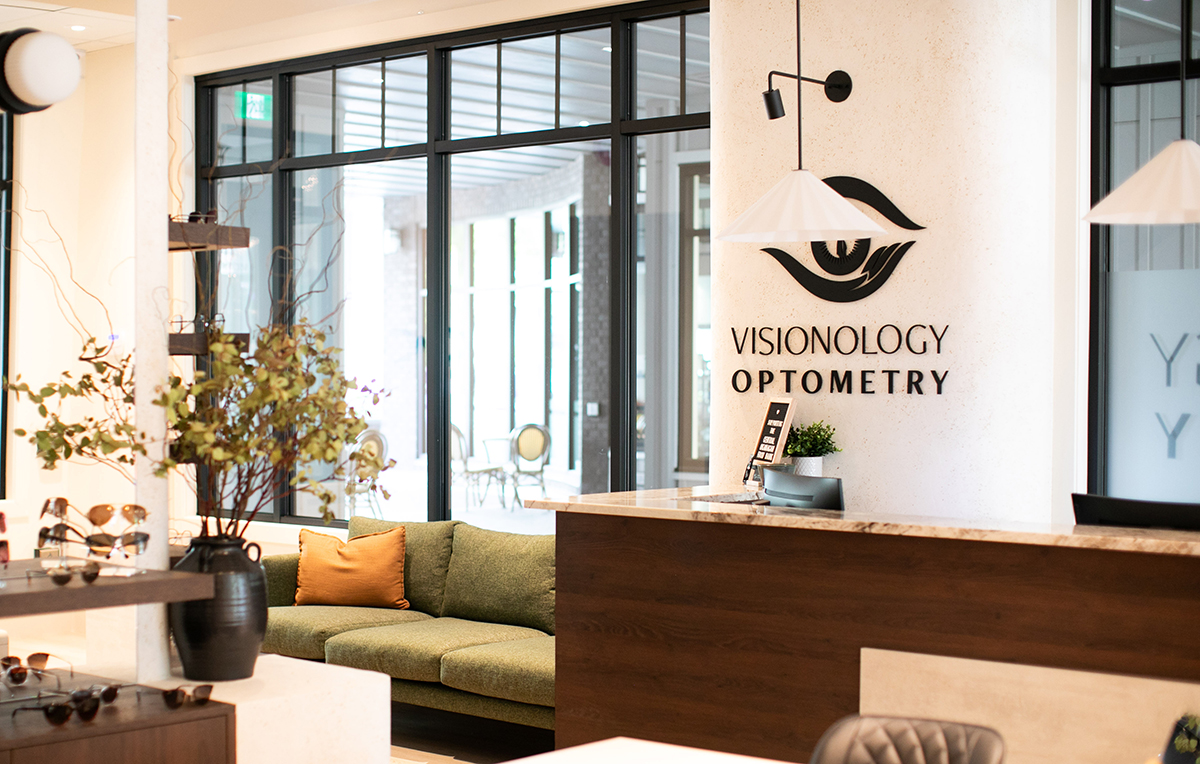 A modern optometry office reception area featuring a sign that reads "VISIONOLOGY OPTOMETRY," a green sofa, decorative plants, and displays of eyeglasses.
