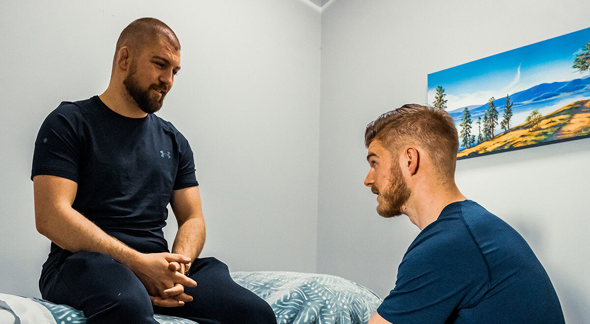 Patient seated on a bed in a room, and therapist with a serious expression while the other, slightly leaning forward, gazes attentively.