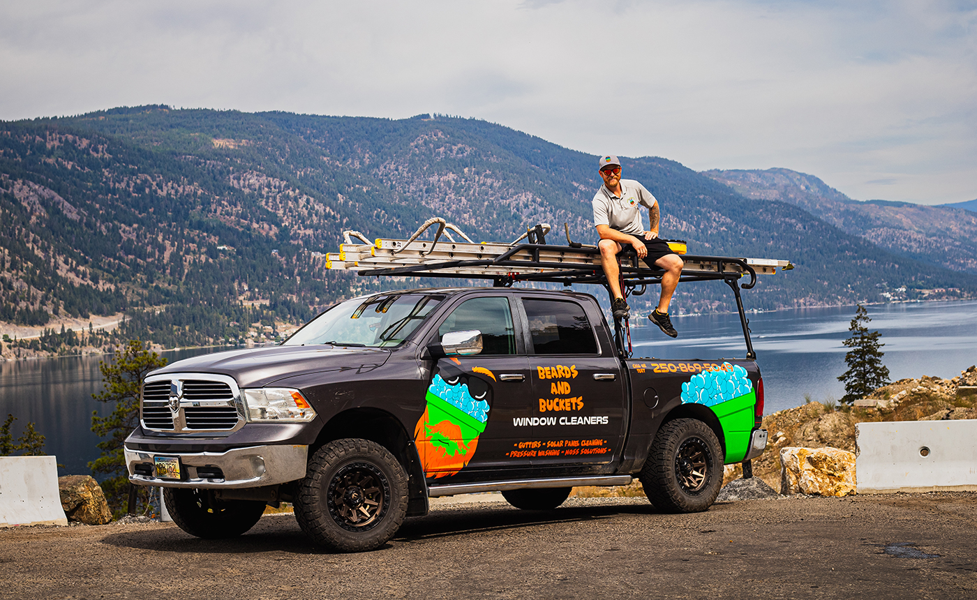A man sits on the roof of a pickup truck equipped with a ladder, parked near a scenic lake surrounded by mountains.