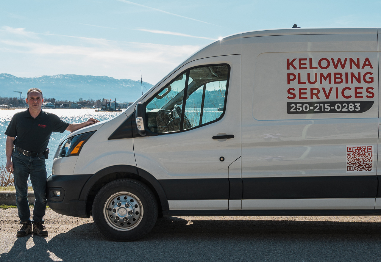 A man stands next to a white van with "Kelowna Plumbing Services" written on the side, by a body of water and mountains in the background.