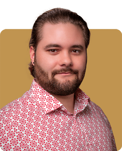 A man with a short beard wearing a patterned pink and white shirt, against a solid background.