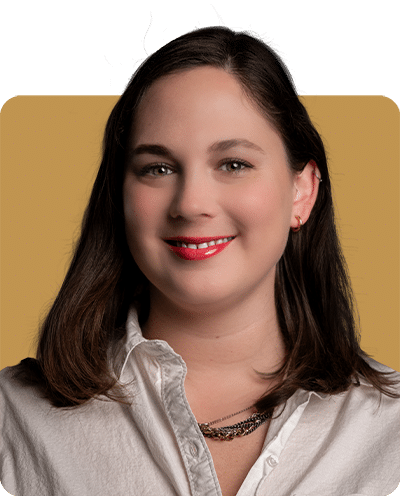 A woman with shoulder-length brown hair, wearing a white shirt and a necklace, smiles against a yellow background.