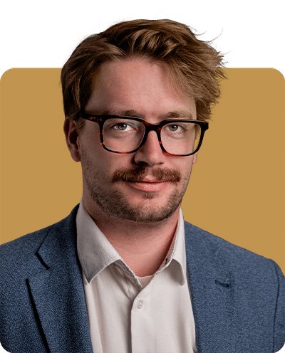A man with glasses and a mustache, wearing a blue blazer and white shirt, against a warm-toned background.