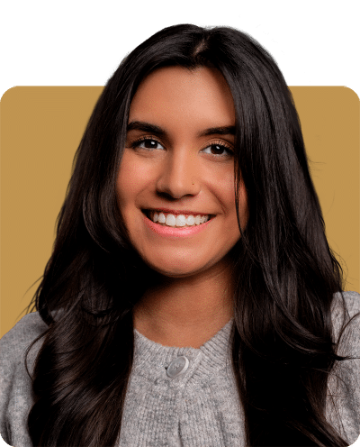 A smiling young woman with long, dark hair, wearing a light gray sweater, against a warm-toned background.