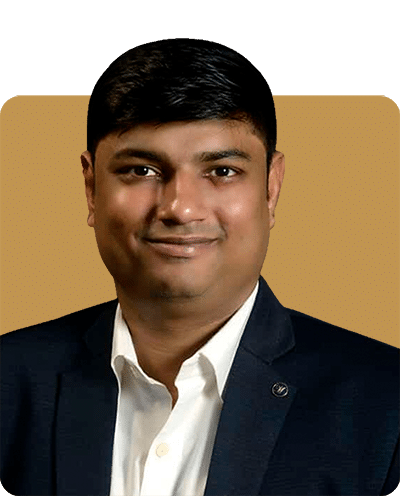 A man smiling, wearing a dark blazer over a white shirt, against a warm-colored background.