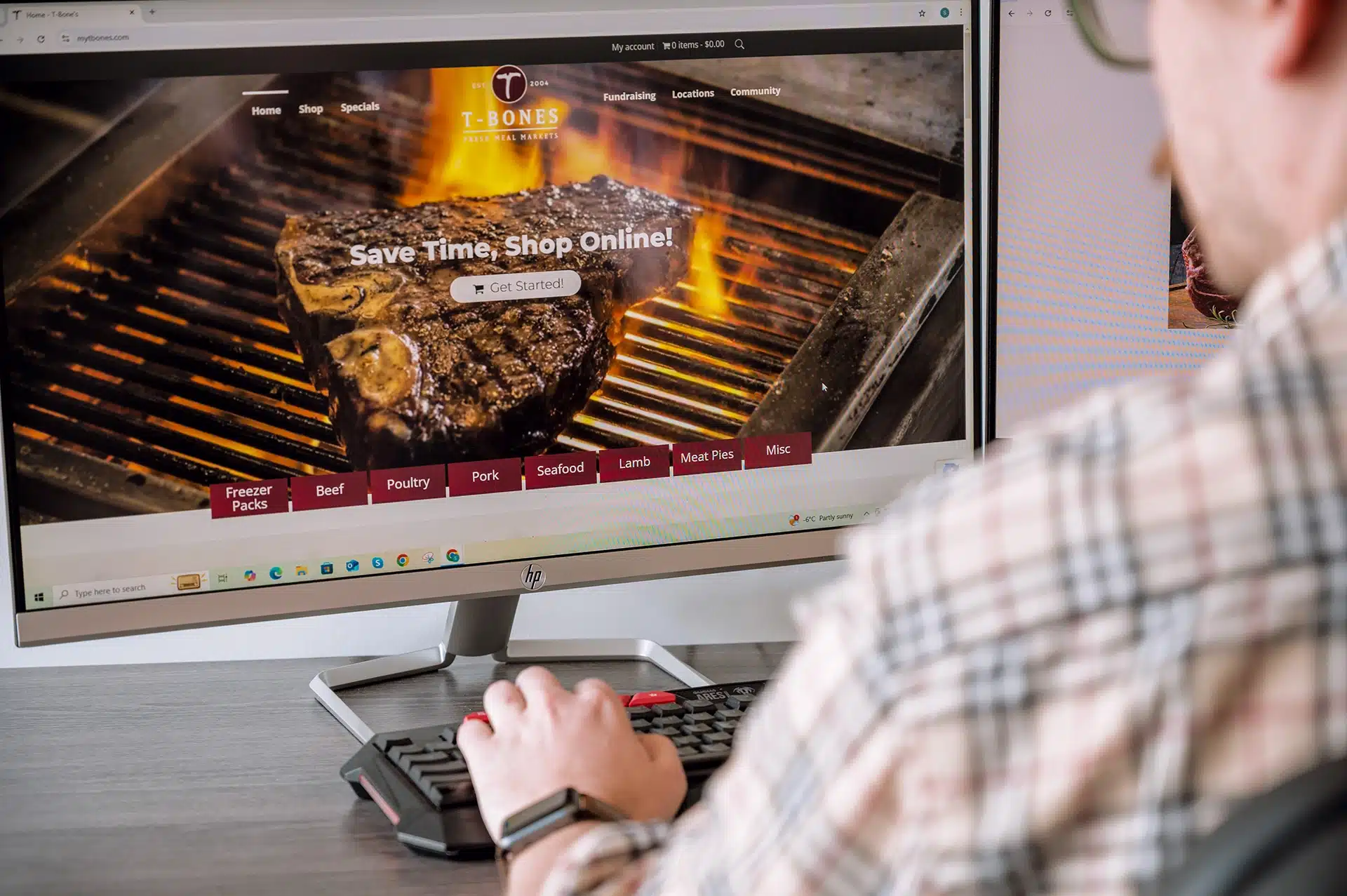 A person is using a computer to view the homepage of T-Bones, a meat market, featuring a grilled steak and shopping options for various types of meat.
