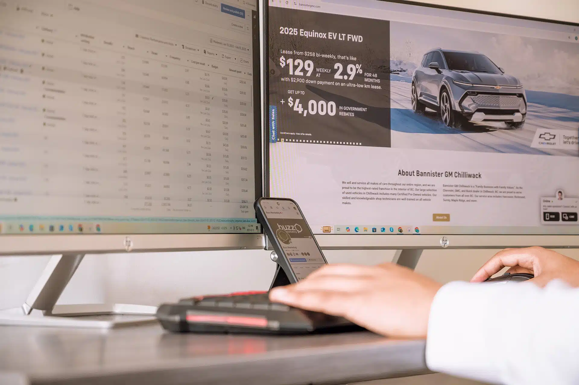 A computer setup with two monitors displaying a car lease advertisement for a 2025 Equinox EV and a spreadsheet. A hand is using a keyboard on the desk.