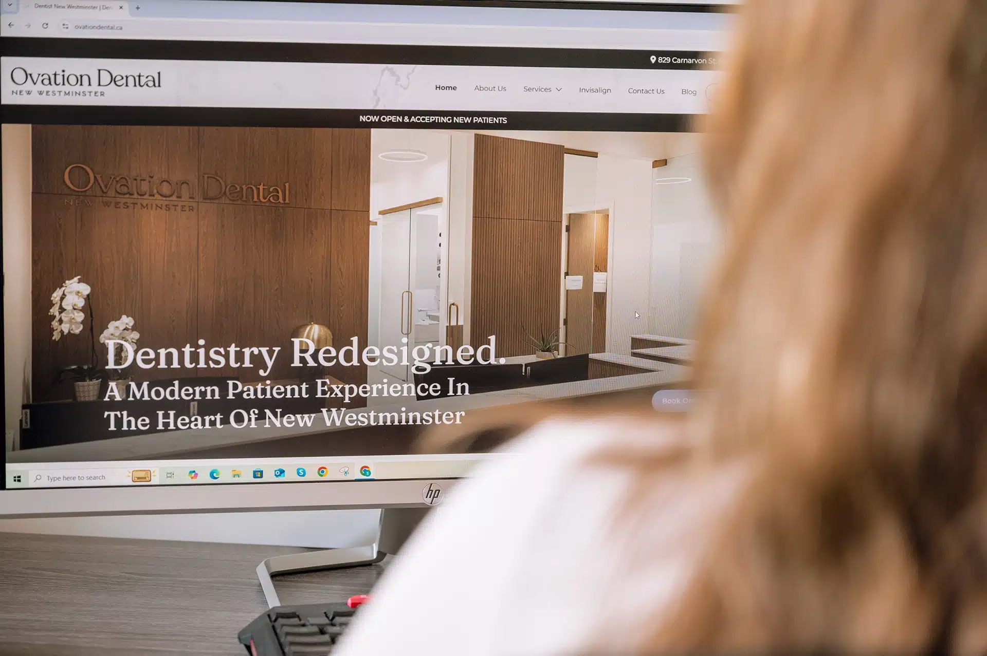 A person is looking at a computer screen displaying the Ovation Dental website, featuring a modern dental office design with the text "Dentistry Redesigned. A Modern Patient Experience In The Heart Of New Westminster."