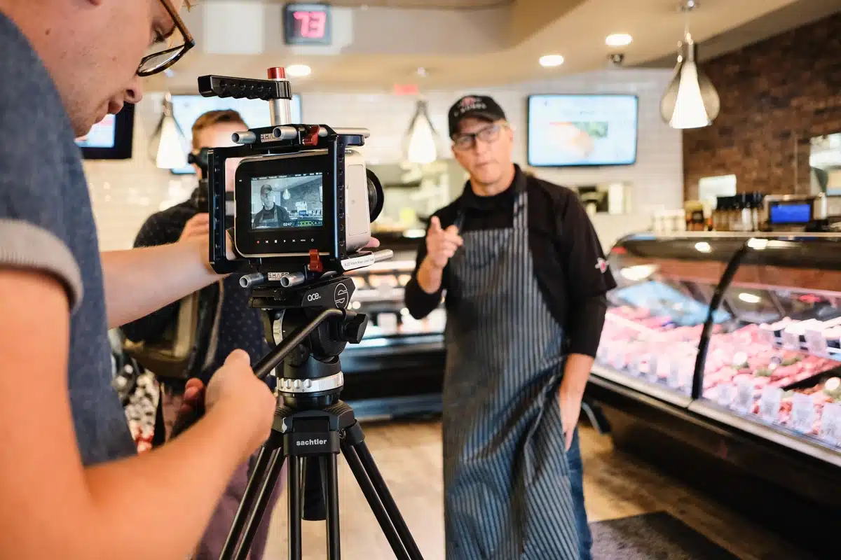 A man in a black shirt and striped apron gestures while being filmed in a market, with a camera setup visible in the foreground. Various meats are displayed in a glass case behind him.