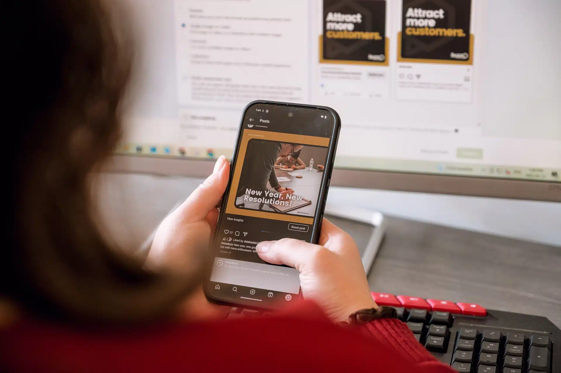A person holding a smartphone displaying a social media post about "New Year, New Resolutions!" in front of a computer screen with online marketing content.