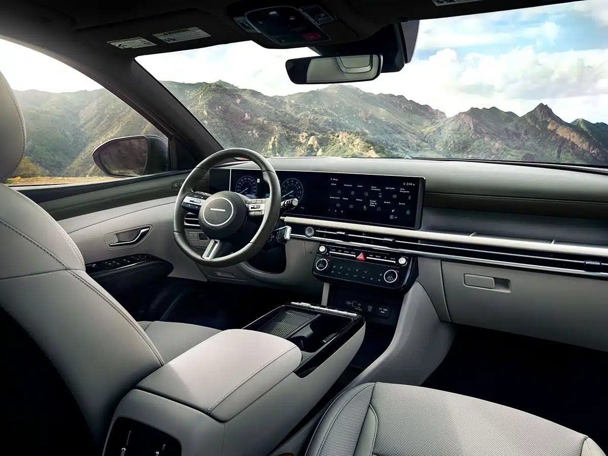 interior of a modern car, highlighting a steering wheel, dashboard with a display screen, and seats in a light gray color, all set against a backdrop of mountains.